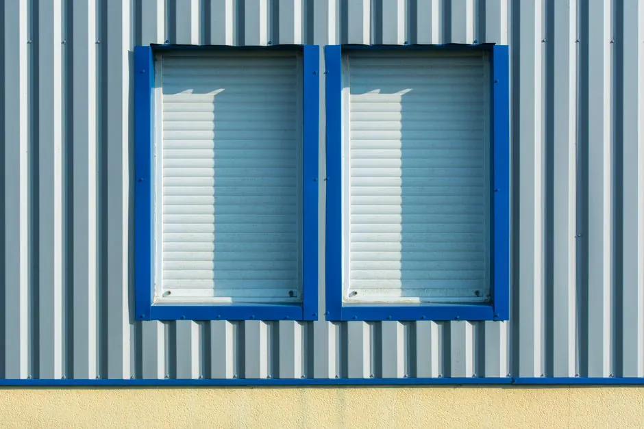 Close-up of corrugated metal wall with blue-framed windows and shutters.